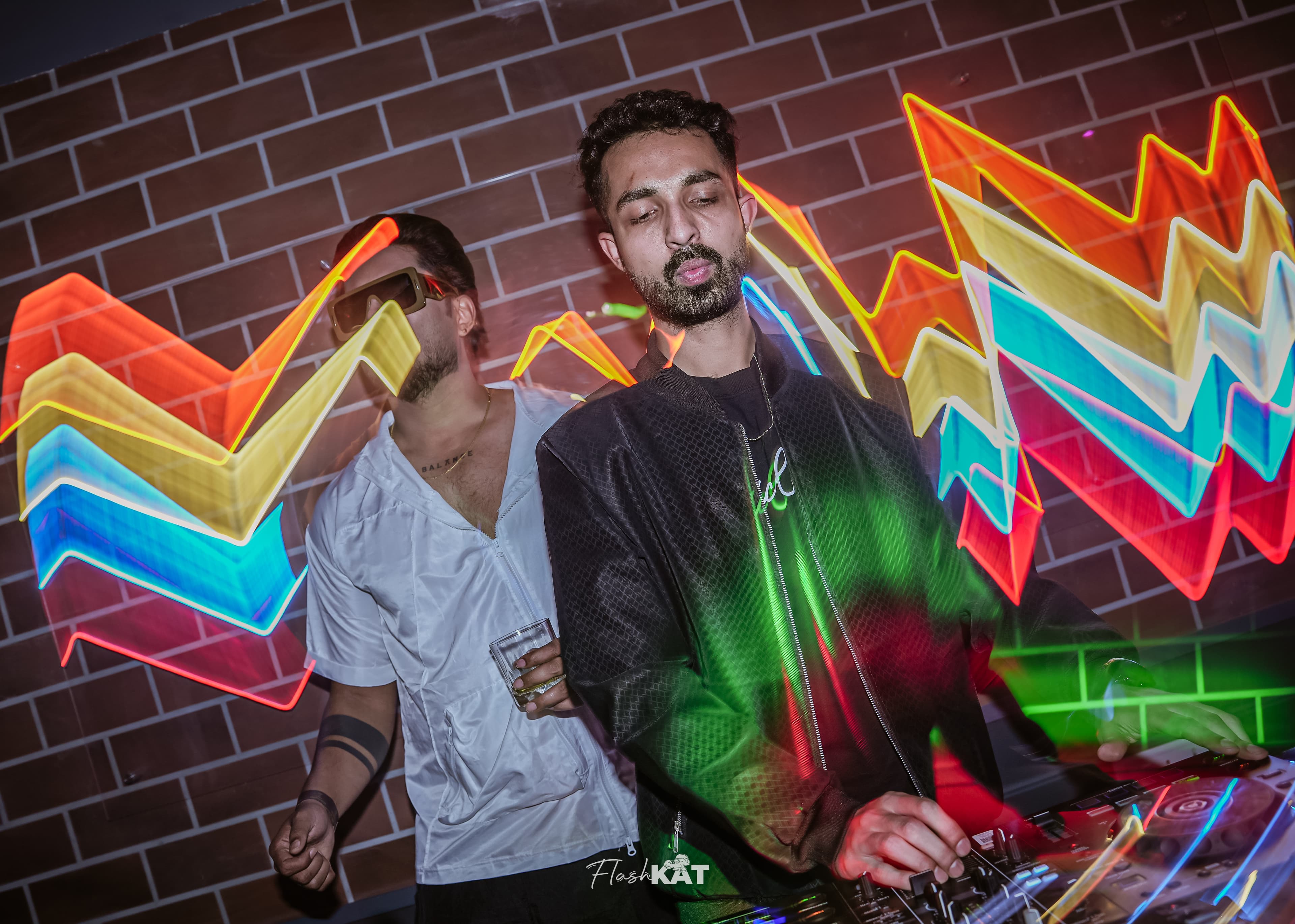 DJ performing at a nightclub with vibrant neon light trails and a brick background.